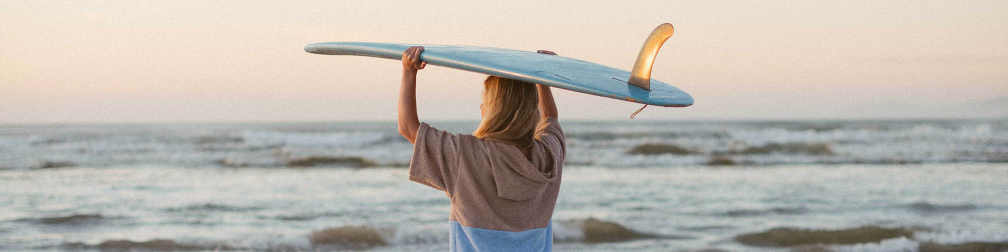 Female holding longboard surfboard in her Mahina salt towelie at sunrise on the beach looking out to the ocean