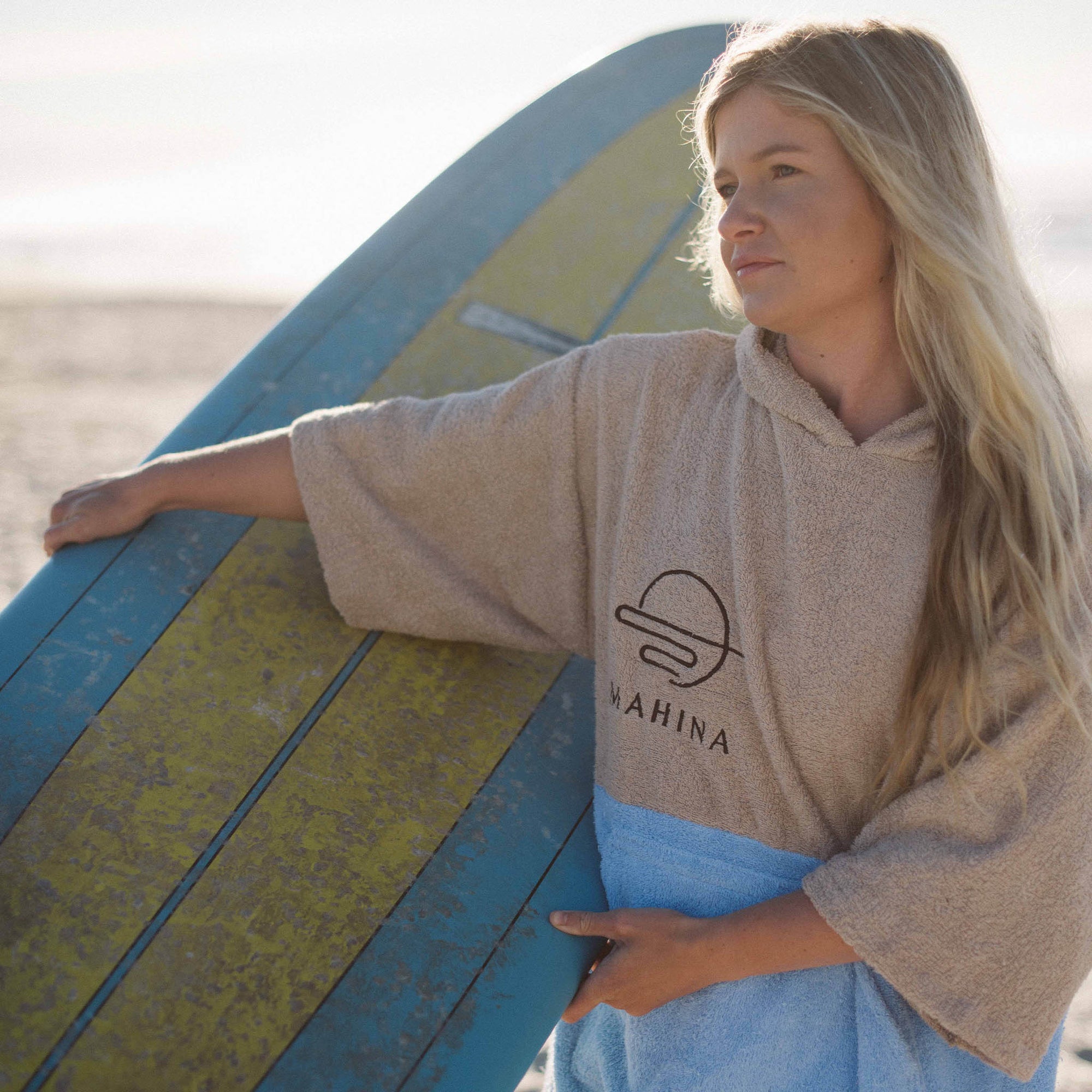 Blonde girl wearing her salt blue Mahina towlie at the beach while holding her surfboard
